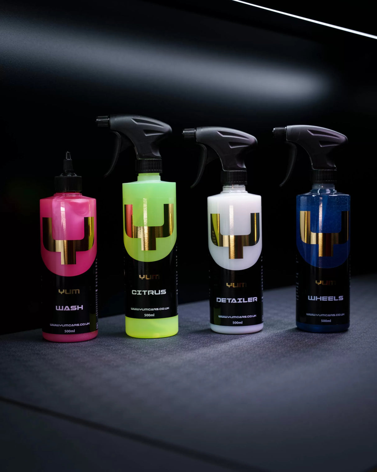 Four YUM car care spray bottles lined up on a dark surface against a black background. The bottles are labeled Wash, Citrus, Detailer, and Wheels, with bright pink, neon yellow-green, white, and blue liquids inside, each featuring a large metallic gold 'V' logo.