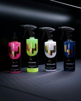 Four YUM car care spray bottles lined up on a dark surface against a black background. The bottles are labeled Wash, Citrus, Detailer, and Wheels, with bright pink, neon yellow-green, white, and blue liquids inside, each featuring a large metallic gold 'V' logo.