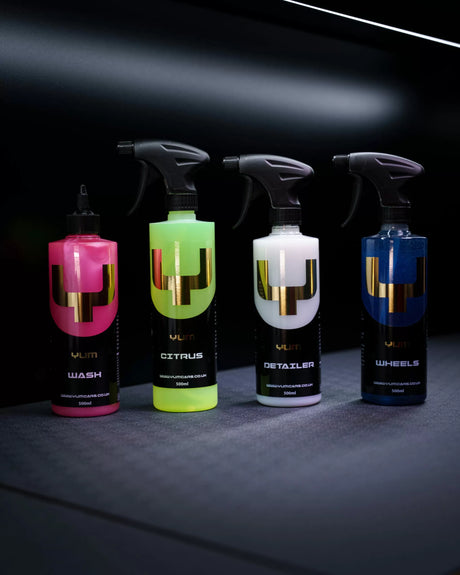 Four YUM car care spray bottles lined up on a dark surface against a black background. The bottles are labeled Wash, Citrus, Detailer, and Wheels, with bright pink, neon yellow-green, white, and blue liquids inside, each featuring a large metallic gold 'V' logo.